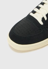 Svart sneakers laget av semsket skinn og mesh, med en perforert tåboks, kremfargede snøringer og en lys gummisåle.
