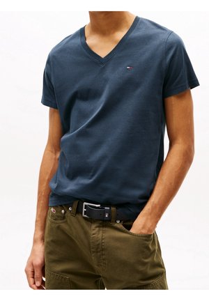 Homme portant un T-shirt bleu marine col en V et un pantalon vert olive avec une ceinture noire, main dans la poche, sur fond blanc uni.