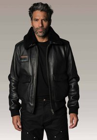 Homme aux cheveux poivre et sel portant une veste en cuir noir avec un col en shearling, une chemise noire et un pantalon noir, se tenant devant un fond uni.
