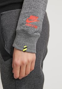 Nike grå sweatshirt med röd logotyp "TRACK & FIELD" på ärmen; mjuk textur, figurnära design och en svart tygdetalj nära handleden.