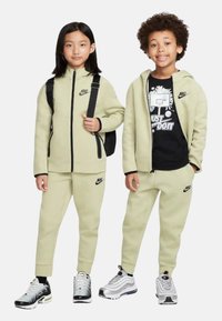 Due bambini indossano felpe con cappuccio e pantaloni della tuta Nike abbinati di colore verde chiaro. Le caratteristiche distintive includono accenti e loghi neri. Il bambino indossa scarpe da ginnastica nere.