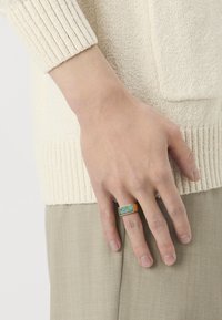 Rektangulær ring med en grønn stein i en gullfarget innstilling. Båret på en hånd, med en lys genser og beige stoff synlig i bakgrunnen.