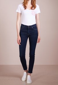 Camiseta blanca de manga corta con escote en V y diseño de flores rosas, combinada con jeans ajustados azul oscuro y zapatillas blancas.