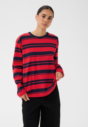 Femme aux cheveux foncés portant un haut à manches longues rouge et bleu marine rayé et un pantalon noir sur un fond clair uni.