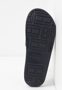 Semelle de sandale en caoutchouc noir avec un motif de grip texturé. Présente un logo et divers motifs rectangulaires. Forme plate et arrondie.