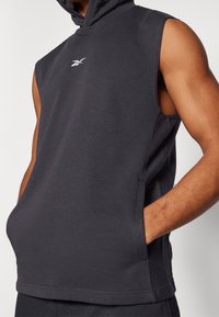Sudadera sin mangas negra con bolsillo canguro, llevada por una persona de pie con una mano en el bolsillo, mostrando un brazo musculoso.