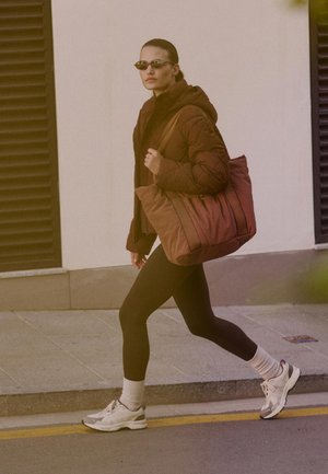 Barna tűzött kabát, fekete leggings, fehér edzőcipő és oversized barna táska. Kiegészítők közé tartozik egy keskeny napszemüveg és fehér zokni.