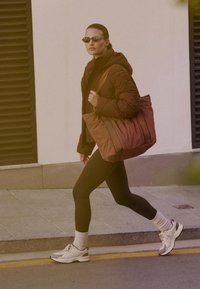Ruske quilted jakke, sorte leggings, hvite sneakers og en overdimensjonert brun tote bag. Tilbehøret inkluderer smale solbriller og hvite sokker.