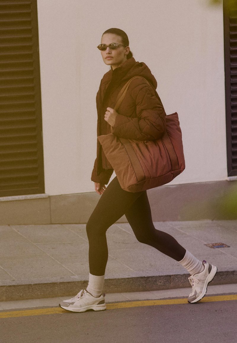 Ruske quilted jakke, sorte leggings, hvite sneakers og en overdimensjonert brun tote bag. Tilbehøret inkluderer smale solbriller og hvite sokker.