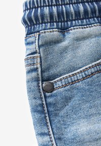 Detailný záber na modré džínsy z denimu so zreteľným prešívaním, kovovým nitom a detailom elastického pásu.