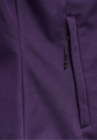 Rip close-up di una tasca di giacca in tessuto viola con una cerniera verticale e un tirante in corda nera sul lato destro.