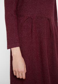 Läheltä otettu kuva henkilön käsivarresta ja kädestä, joka on pukeutunut pitkähihaiseen, tummanpunaiseen neulemekkoon, jossa on pleated-yksityiskohtia vyötäröllä.