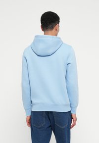 Άντρας που φοράει ανοιχτό γαλάζιο φούτερ και μ�πλε τζιν, απεικονιζόμενος από πίσω σε λευκό φόντο.