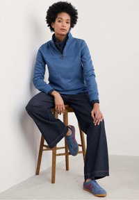 Pullover in pile blu con collo a zip, jeans larghi scuri e sneakers blu con accenti rossi, seduto su uno sgabello di legno davanti a una parete semplice.