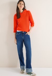Femme portant un sweat à capuche orange vif, un jean bleu retroussé et des baskets blanches avec des rayures vertes, debout de manière détendue contre un mur clair.