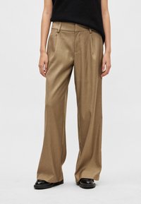 Szerokie spodnie z lekkiego brązowego materiału o gładkiej fakturze, wyposażone w boczne kieszenie oraz wysoki stan. W połączeniu z czarnymi butami.