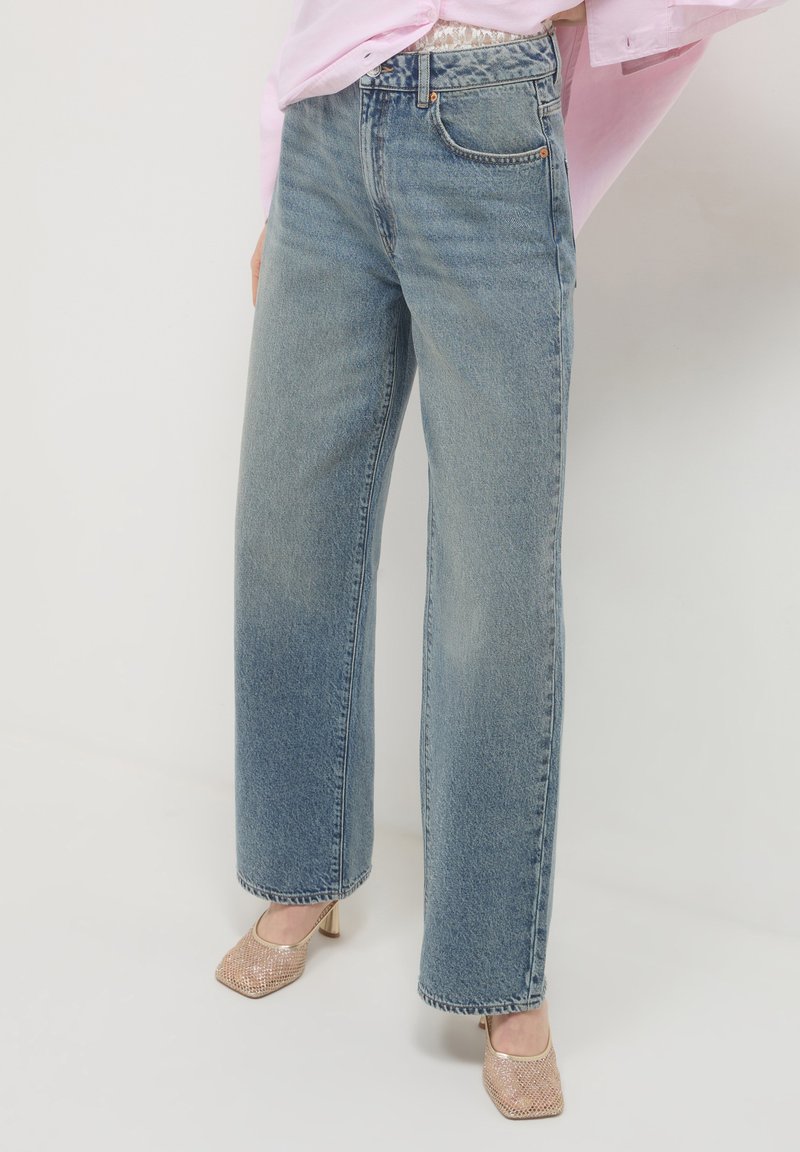 Donna che indossa jeans blu a vita alta e gamba larga abbinati a scarpe con tacchi alti dorate e una blusa rosa.