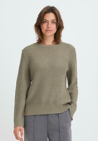 Khaki-Strickpullover mit einem strukturierten Muster, Rundhalsausschnitt und geripptem Saum; getragen mit grauen Hosen.