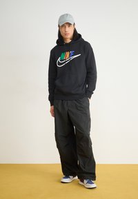 Felpa nera con logo Nike multicolore, pantaloni cargo neri e cappellino grigio. Le calzature presentano accenti blu, bianchi e rossi. Texture lisce.