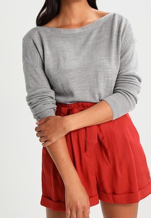 Vrouw draagt een lichtgrijze top met lange mouwen met geplooide mouwen en hooggetailleerde rode shorts, vastgebonden met een stoffen riem.