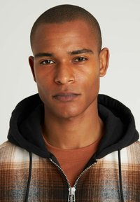 Sudadera con capucha de un patrón de cuadros marrón y crema, con capucha negra y cierre de cremallera, llevada sobre una camiseta de color óxido.