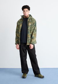 Πράσινο φλις μπουφάν με καμουφλάζ και φερμουάρ, μαύρη μπλούζα, μαύρο παντελόνι και πράσινα παπούτσια πεζοπορίας. Το μπουφάν έχει λογότυπο στο στήθος.