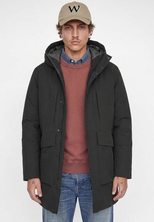 Homme portant une casquette beige, un manteau d’hiver noir à capuche sur un pull couleur rouille et un jean bleu, debout devant un fond blanc uni.