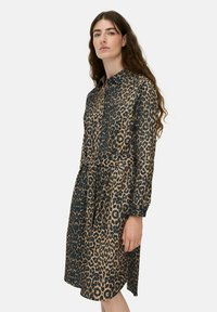 Frau mit langen braunen Haaren, die ein knielanges Hemdkleid mit Leopardenmuster, langen Ärmeln und einem Taillengurt trägt, steht vor einem weißen Hintergrund.
