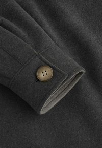 Tessuto in lana grigio scuro con una texture morbida, caratterizzato da una chiusura a bottone al polsino e sottili dettagli di cucitura.