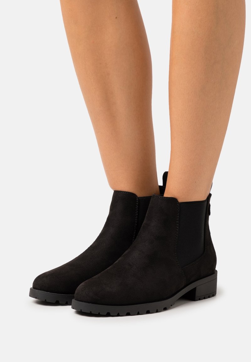 Bottines en daim noir avec panneaux latéraux élastiques et talons carrés bas portées sur des jambes nues sur un fond uni.