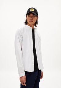 Witte overhemd met een bandkraag, gecombineerd met een zwart T-shirt, donkere spijkerbroek en een zwarte pet met een geel logo.