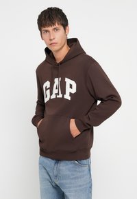 Brown hoodie made of cotton blend with white "GAP" lettering. Features a kangaroo pocket and adjustable drawstring hood. Worn with light blue jeans.