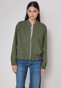 Giacca bomber verde oliva con zip, polsini elasticizzati e orlo con cordoncino. Presenta un tessuto liscio e due tasche laterali.