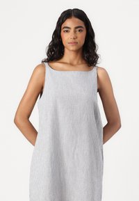 Rochie fără mâneci, în dungi gri și albe, cu decolteu pătrat, buzunare laterale și o croială lejeră. Textură de țesătură moale și ușoară.