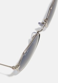 Metalizuoti sidabriniai akiniai nuo saulės su tamsiai pilkais lęšiais, unikalios formos, reguliuojamais nosies pagalvėlėmis ir detalizuotais šonų akcentais. Matoma Ray-Ban prekės ženklas.