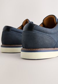 Paire de chaussures décontractées en daim bleu marine avec semelles blanches et coutures marron, vues de dos sur fond blanc.