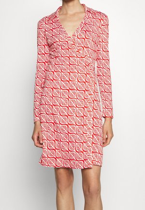 Wikkeljurk met lange mouwen en rood-wit bladerenpatroon, tot de knie reikend, gedragen door een persoon die tegen een effen achtergrond staat.