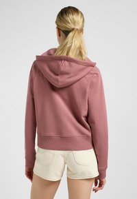 Sudadera corta rosa con capucha ajustable, puños y dobladillo acanalados. Combinada con pantalones cortos de color claro, mostrando un ajuste relajado y una textura suave.