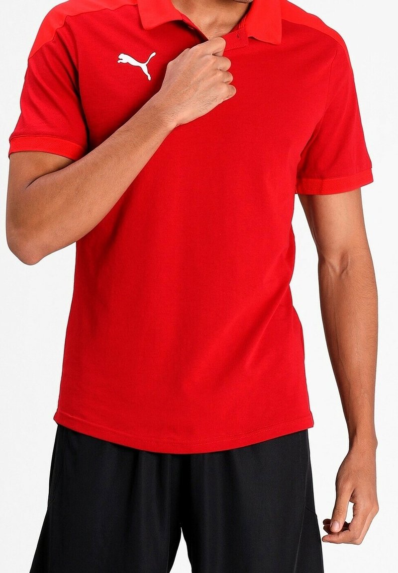 Homme portant un polo rouge Puma, ajustant le col, associé à un short noir, sur fond clair.