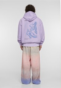 Lavandas krāsas kapucei uz muguras ir abstrakts zils raksts. Kombinēta ar platām biksēm, kuru krāsu gradients svārstās no rozā līdz zila toņiem. Audums ar tekstūru.