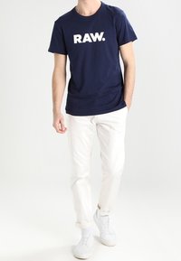 Marinblå bomullströja med en stor vit "RAW."-logotyp, tillsammans med vita byxor och vita sneakers, stående mot en ljus bakgrund.