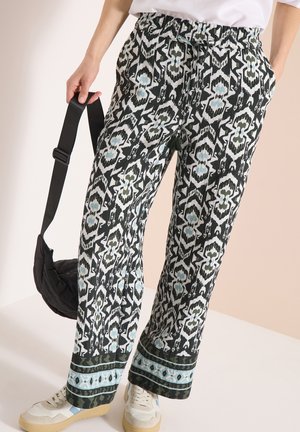 Personne portant un pantalon ample à motif noir et blanc avec des accents bleus, des baskets blanches, tenant un petit sac à bandoulière noir.