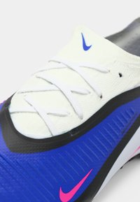 Αθλητικό παπούτσι με λευκό επάνω μέρος με μπλε λεπτομέρειες, μαύρη επικάλυψη και ροζ λογότυπο Nike. Οι κορδόνια είναι λευκά· ο υφασμένος πάτος είναι ορατός.