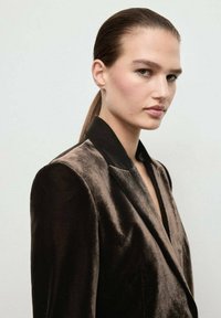 Joven mujer con el cabello peinado hacia atrás, vistiendo un blazer de terciopelo oscuro, mirando ligeramente hacia un lado contra un fondo claro y liso.