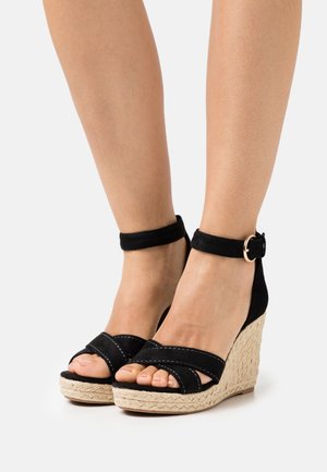 Sandalias cuñas negras con plataforma de fibra de yute. Presentan tiras de ante cruzadas y una correa en el tobillo con una hebilla dorada. Textura suave.