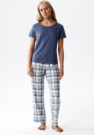 Młoda kobieta stojąca boso, ubrana w granatową koszulkę z krótkim rękawem oraz niebiesko-biało-brązowe, kraciaste spodnie do piżamy na białym tle.