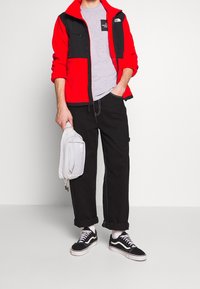 Osoba ubrana w czerwono-czarną kurtkę na zamek, szary T-shirt, czarne spodnie, czarne sneakersy, trzymając małą jasnoszara torbę za uchwyt.