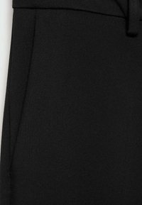 Gros plan d’un pantalon noir sur mesure montrant la poche latérale, la taille et le passant de ceinture sur un tissu texturé.