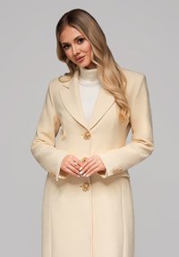 Cappotto su misura color crema con colletto a punta, chiusura con bottoni dorati e maniche lunghe. Indossato sopra un maglione a collo alto bianco.