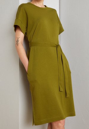 Woman wearing knee-length olive green dress with short sleeves, fabric belt, and side pockets, standing against a neutral wall.